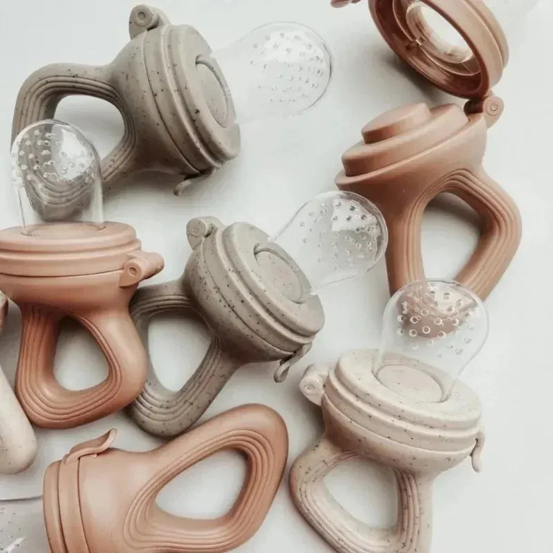 Assorted silicone baby fruit feeders in speckled beige and solid peach, with soft clear nipples.