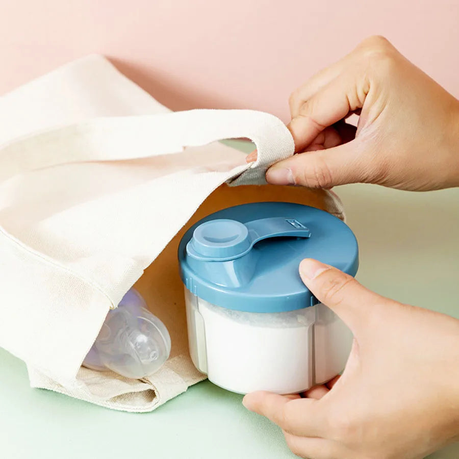 Portable 4-compartment baby formula dispenser with blue lid, being placed into a canvas bag next to a baby bottle.
