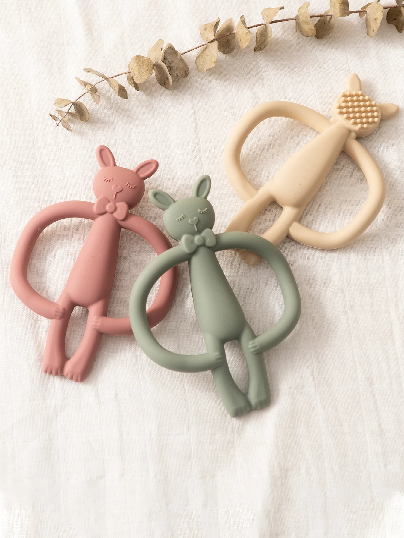 Three soft silicone bunny teethers in pink, green, and beige, with an ergonomic ring design.