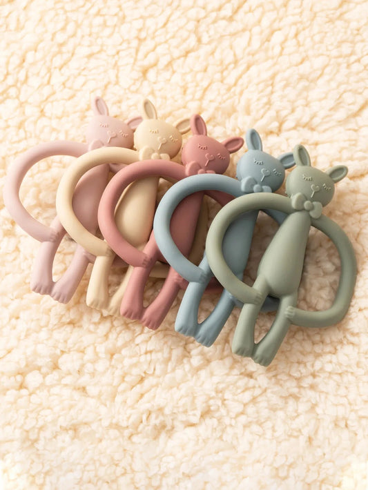 Soft silicone bunny teethers in pastel pink, yellow, peach, blue, and green on a cream fuzzy blanket.