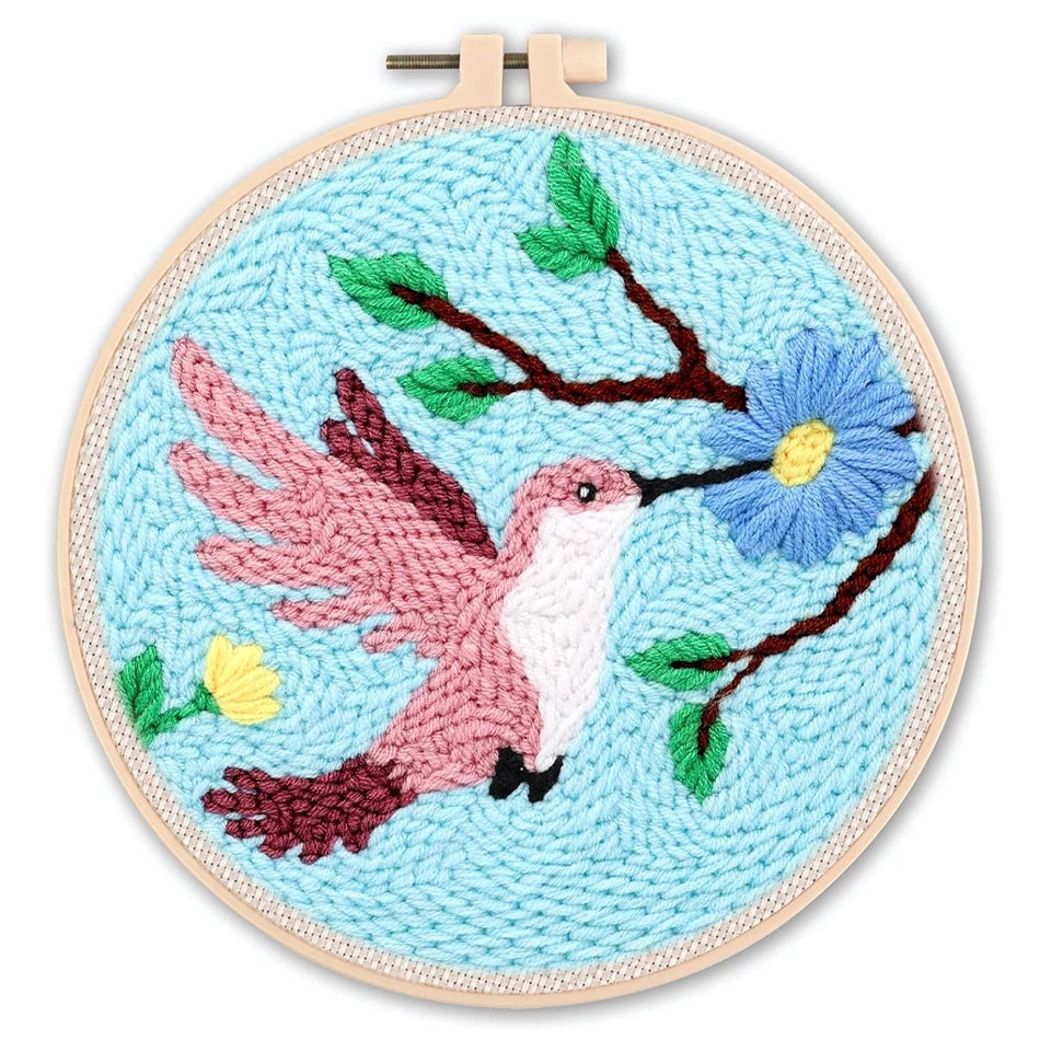 Punch needle embroidery kit featuring a pink hummingbird, blue flower, and green leaves on a light blue background in a wooden hoop.