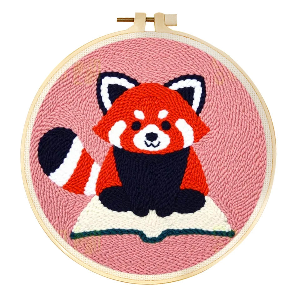 Punch needle embroidery kit featuring a cute red panda on a book, set in a wooden hoop with pink background.