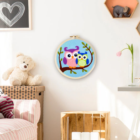 Punch needle embroidery kit with two colorful owls on a branch in a wooden hoop, hanging on a white wall.