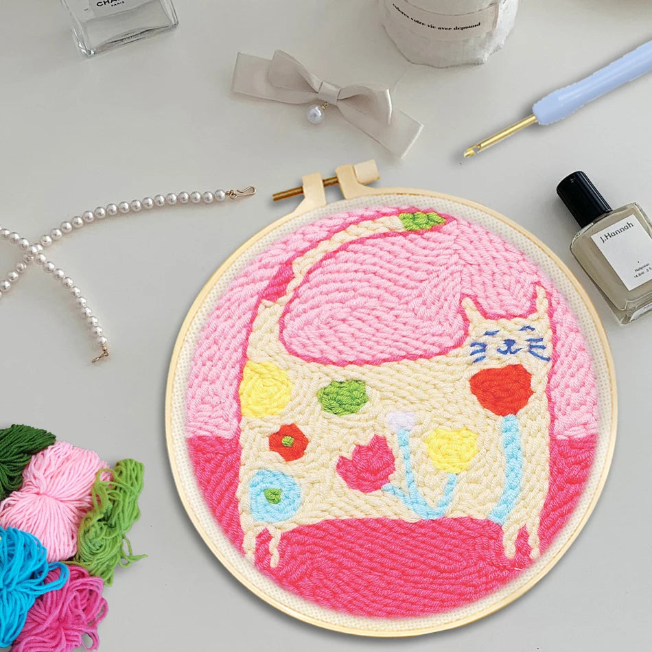 Punch needle embroidery kit with a cute pink cat design in a wooden hoop, surrounded by colorful yarn.
