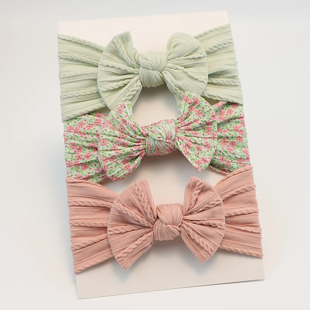 Set of 3 soft nylon baby headbands: light green ribbed, pink floral, and pink ribbed with bows.