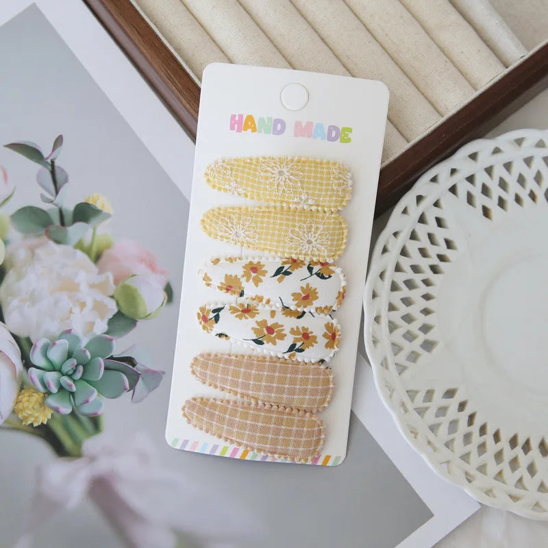 Handmade set of 6 fabric snap hair clips for girls, featuring yellow floral, checkered, and embroidered designs.