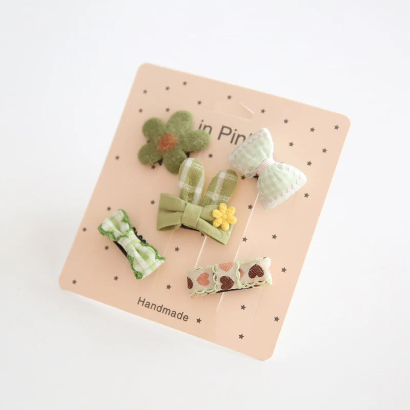 Set of five green and white baby hair clips with bows, bunny ears, flower, and heart designs on a pink card.