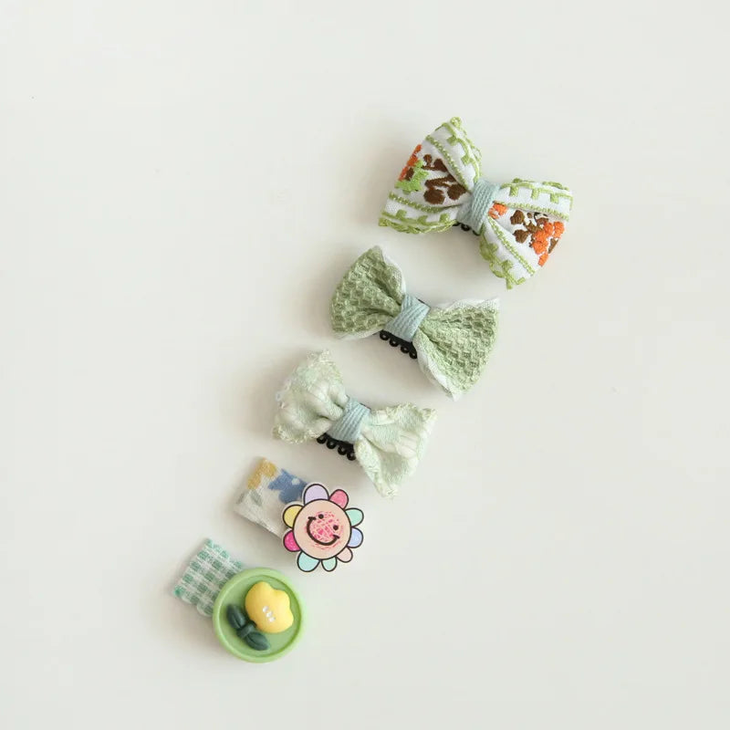 Set of five baby hair clips: two embroidered floral bows, two textured green bows, and one clip with a smiling flower.