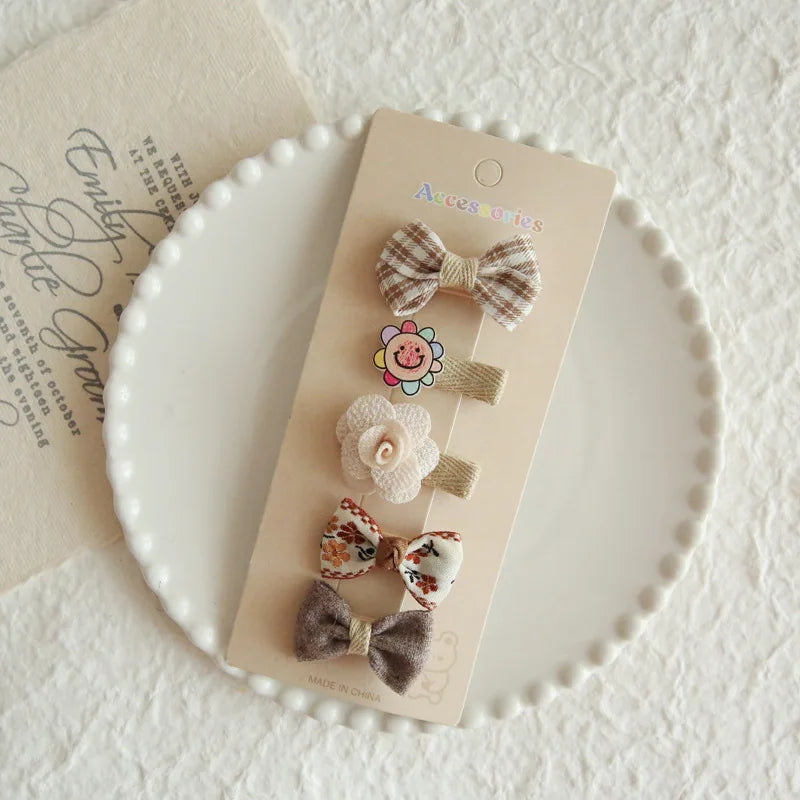 Set of 5 baby girl hair clips with floral, bow, and smiley face designs in neutral colors.