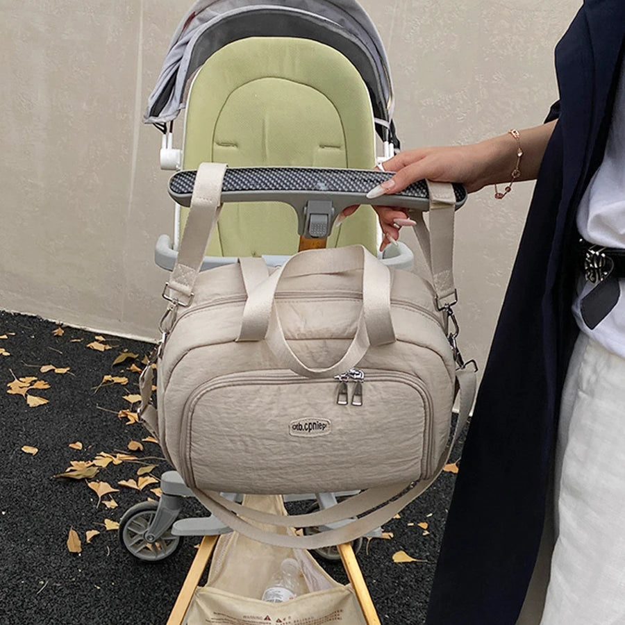 Beige multi-functional diaper bag with shoulder strap, hanging on a stroller, held by a woman.