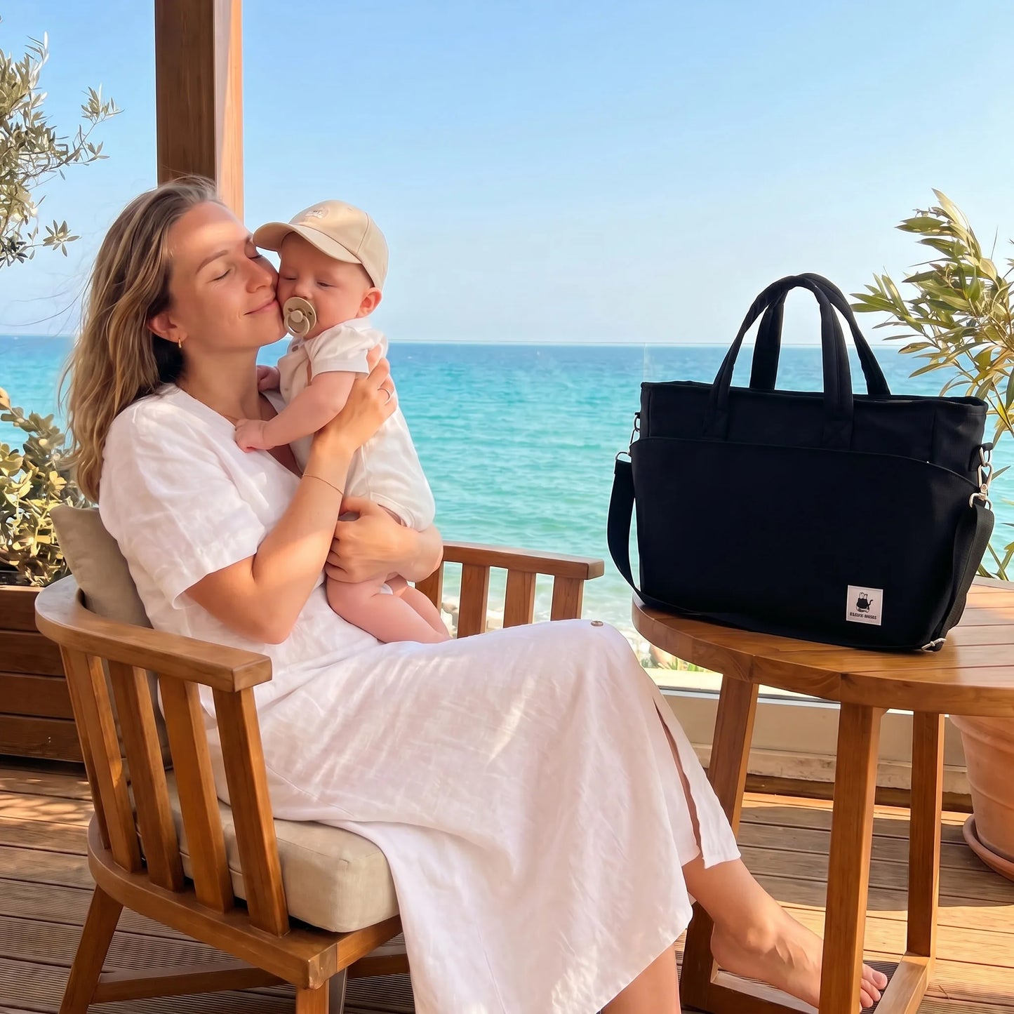 Black insulated diaper bag on a wooden table next to a mother holding her baby by the sea.