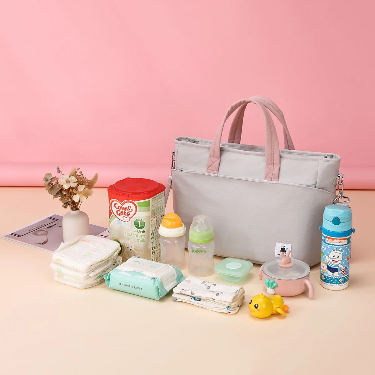 Gray insulated diaper bag with pink handles, surrounded by baby bottles, formula, diapers, and wipes.