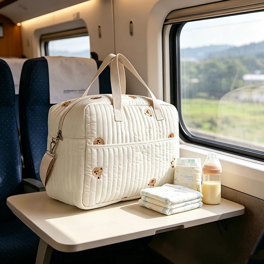 Cream quilted polyester diaper bag with teddy bear embroidery, baby bottle, and diapers on a train table.