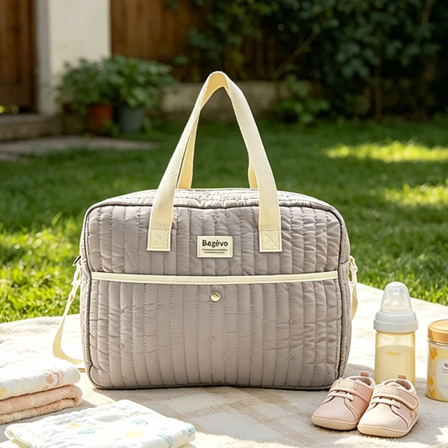 Quilted grey diaper bag with cream straps, baby bottle, pink shoes, and blankets on a picnic blanket outdoors.