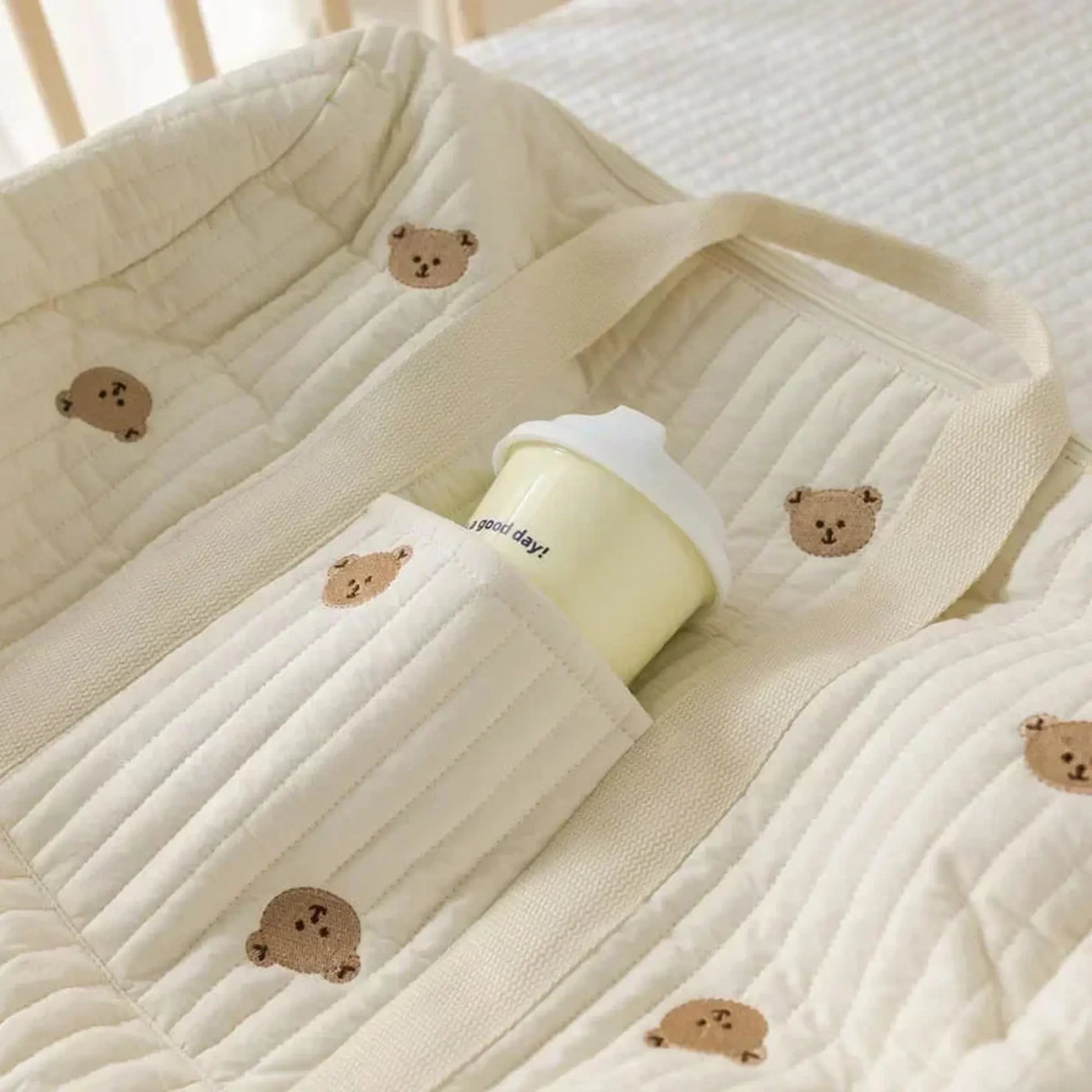 Cream quilted cotton diaper bag with embroidered teddy bears and a bottle in a side pocket.