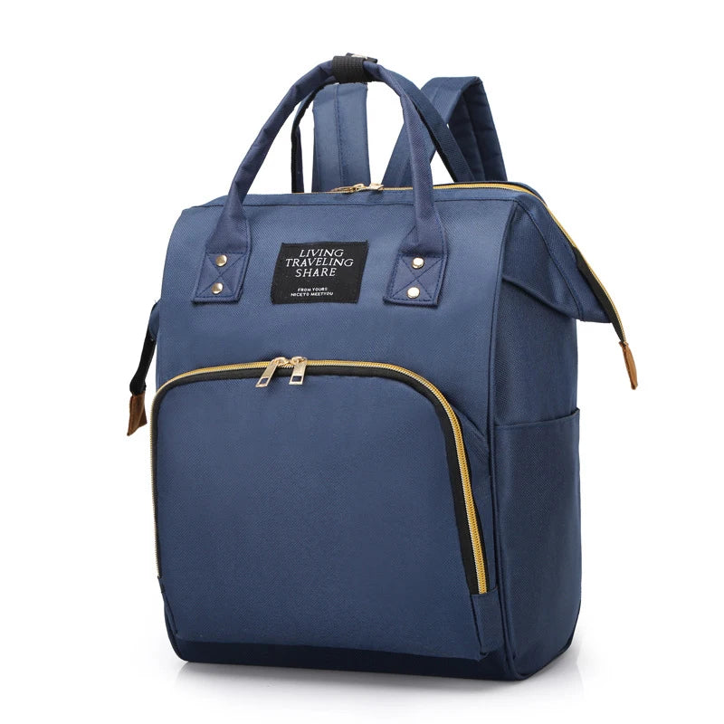 Navy canvas diaper backpack with gold zippers, top handles, and "LIVING TRAVELING SHARE" patch.